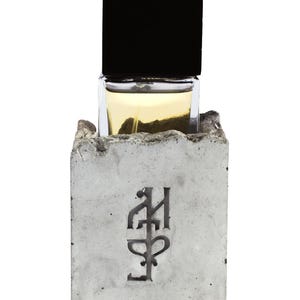 May include: A rectangular glass perfume bottle with a black cap, partially encased in a rough, grey concrete block. The concrete base features a dark, stylized engraved symbol on the front face, and the bottle contains a pale yellow liquid.
