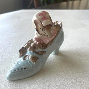 May include: A ceramic figurine of a mouse wearing a pink and white bonnet, seated inside a light blue high-heeled shoe. The shoe has brown accents and decorative cutouts. The figurine is a whimsical decorative item.