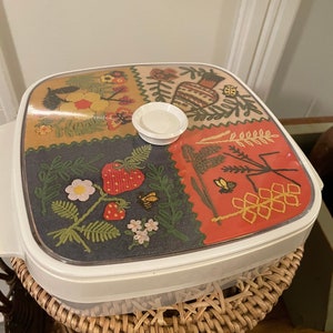 May include: A vintage white and clear plastic serving dish with a decorative lid. The lid features a colorful patchwork design with floral, strawberry, and bee motifs. The dish is sitting on a woven wicker basket.