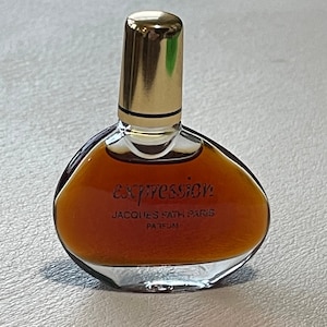 May include: A small, clear glass perfume bottle with a gold-colored cap. The bottle contains a brown liquid and has the word "expression" printed on the front, along with "JACQUES FATH PARIS PARFUM".