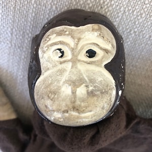 May include: A hand puppet featuring a monkey head with a dark brown painted surface and a light cream-colored face. The monkey has black circular eyes and a sculpted nose and mouth area. The puppet is attached to a dark brown fabric body.