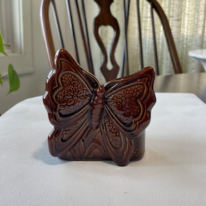 May include: A brown ceramic butterfly-shaped napkin holder. The butterfly has detailed wing patterns and a glossy finish. The holder is designed to stand upright and is likely for decorative use on a dining table or kitchen counter.