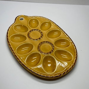 May include: A tan ceramic egg serving platter with 12 oval indentations. The platter has a decorative rope-like border and a small hole for hanging. The center and border are accented with a dark brown color.