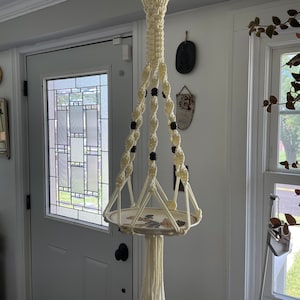 May include: Cream-colored macrame plant hanger with a decorative plate. The plant holder features a woven design with dark beads and a circular plate with a floral pattern. The hanger is suspended in front of a white door with a stained glass window.