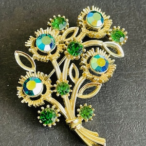 May include: A gold-toned floral brooch featuring a bouquet design. The brooch has green and iridescent blue-green rhinestones. The flowers are surrounded by gold-toned petals and leaves. The brooch is a vintage style.