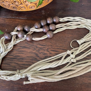 May include: A beige macrame plant hanger with dark brown wooden beads. The plant hanger is made of woven rope and has a metal ring for hanging. The design is suitable for a small to medium-sized plant pot.