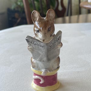 May include: A figurine of a brown mouse reading a newspaper titled "The Tailor Cutter." The mouse is perched atop a spool of thread with a red center, yellow base, and a white thread detail. The figurine has a detailed design.