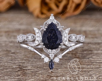 May include: A pear-shaped blue starry night gemstone engagement ring set with a delicate, intricate design featuring crescent moons and floral motifs. The ring is set in silver and features a matching band.