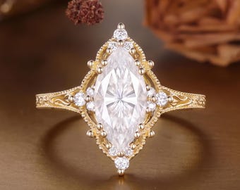 May include: A gold ring featuring a large marquise-cut center stone surrounded by a halo of smaller round stones. The band displays intricate metalwork details, and the setting has a pointed, elongated shape with decorative milgrain edges.