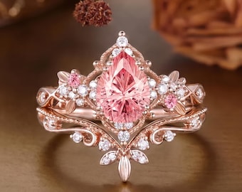 May include: A rose gold ring set featuring a pear-shaped peach-colored center stone, accented with small round white diamonds and two small pink stones. The band and setting display intricate metalwork with floral-inspired filigree details.