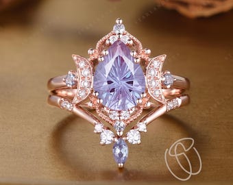 May include: A rose gold ring featuring a pear-shaped lavender center stone surrounded by a halo of small white round stones. The band includes crescent moon motifs on each side and a pointed V-shaped design adorned with additional white stones.