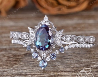 May include: A pear-shaped alexandrite engagement ring set with a delicate, intricate design featuring two crescent moons and a halo of smaller diamonds. The ring is set on a white gold band and is accompanied by a matching wedding band with a row of diamonds.