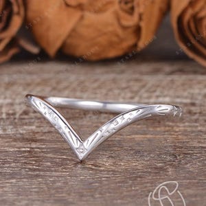 May include: A silver V-shaped ring with etched detailing. The ring has a chevron design and is set against a wooden surface with dried flowers in the background. The ring is a delicate piece of jewelry.
