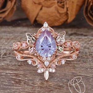 May include: A rose gold engagement ring set with a pear-shaped, light purple gemstone. The ring features intricate filigree and small accent stones. The image also shows the text "STARLANDUS".