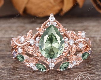 Puede incluir: Un anillo de piedra preciosa verde en forma de pera con un delicado diseño floral y piedras preciosas verdes más pequeñas. El anillo está engastado en oro rosa y presenta un halo de diamantes más pequeños.