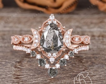 Puede incluir: Un anillo de compromiso de diamante gris en forma de pera engastado en oro rosa con un diseño delicado e intrincado. El anillo presenta un motivo de luna creciente y un halo de diamantes más pequeños. La banda está adornada con un delicado patrón de filigrana.