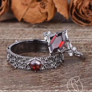 May include: Two rings with a gothic design. One ring features a large, faceted, red gemstone in a unique setting. The other ring has a matching red gemstone and intricate filigree detailing. The rings are set against a wooden background.