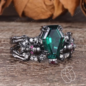 May include: A black gothic ring featuring a coffin-shaped, emerald-green gemstone. The ring has a detailed design with small, round pink and white gemstones, and a dark, skeletal-like structure. The ring is set against a wooden surface.