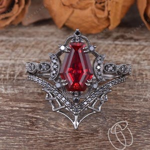 Peut inclure: Une bague argentée avec une grosse pierre rouge facettée au centre. La bague présente des détails complexes avec de plus petites pierres noires et un design décoratif d'inspiration gothique. La bague est posée sur un fond en bois.