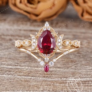 Puede incluir: Un anillo de rubí en forma de pera con una banda de oro delicada y una banda a juego con un diseño de luna creciente. El anillo está engastado con diamantes y rubíes más pequeños.