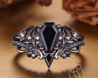 May include: A black metal ring featuring a central coffin-shaped black gemstone, flanked by intricate filigree metalwork and smaller black stones. The band includes a curved design with additional black stones set along the sides and a small pointed accent.