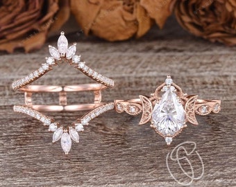 May include: A pear-shaped diamond engagement ring with a delicate floral design and two crescent moons on either side of the center stone. The ring is set in rose gold and is accompanied by a matching rose gold wedding band with a curved design and a row of marquise-shaped diamonds.