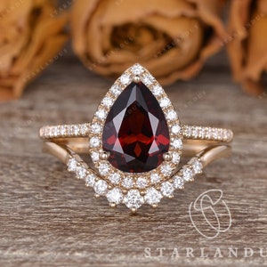 May include: A pear-shaped garnet engagement ring with a halo of smaller diamonds set in a gold band. The ring is paired with a matching wedding band with a curved design and smaller diamonds.