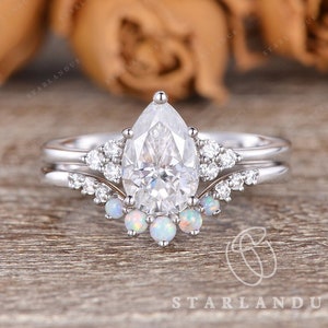 May include: A pear-shaped diamond engagement ring with a delicate band and a halo of smaller diamonds. The ring is set with a row of opal stones below the center stone.