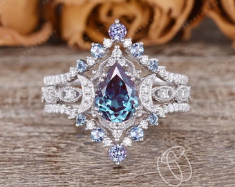 May include: A pear-shaped alexandrite engagement ring with a halo of smaller diamonds and blue sapphires. The ring is set in a white gold band and features a delicate, intricate design.