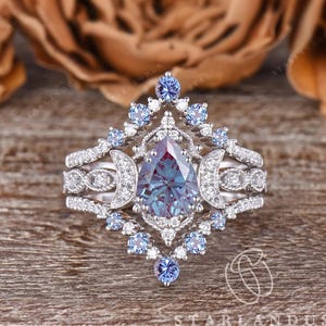 Könnte beinhalten: Ein dreiteiliges silbernes Ring-Set mit einem birnenförmigen, farbwechselnden Edelstein. Der Ring ist von kleinen blauen Edelsteinen und weißen Diamanten umgeben. Das Set beinhaltet ein Band mit einer Reihe von Diamanten und zwei Mond-Designs.