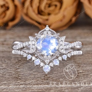 May include: A silver engagement ring set with a large round moonstone center stone surrounded by smaller diamonds. The ring has a delicate filigree design and a crescent moon detail on each side of the center stone. The band is a twisted design with smaller diamonds set along the band.