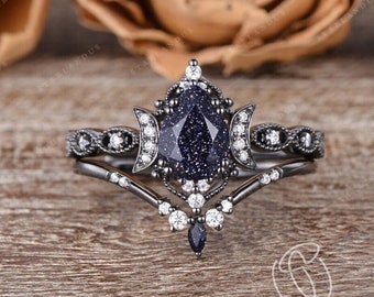 Può includere: Un set di anello di fidanzamento in oro nero con una pietra preziosa blu a forma di pera circondata da pietre bianche più piccole. L'anello presenta un design a forma di luna crescente e dettagli delicati.