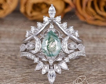 Puede incluir: Un anillo de plata con una piedra preciosa de ágata verde musgo en forma de pera, rodeada de pequeñas piedras preciosas transparentes. El anillo presenta un diseño delicado e intrincado con un motivo de luna creciente.