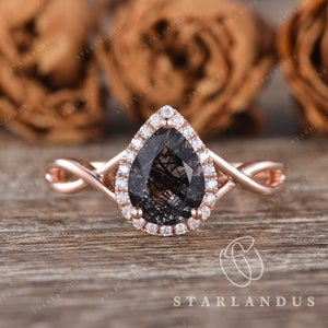 May include: A rose gold engagement ring with a pear-shaped black diamond center stone surrounded by a halo of smaller diamonds. The band is a twisted design.