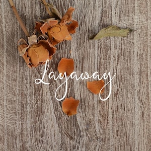 May include: A close-up shot of a wooden surface with the word "Layaway" in elegant cursive script. Dried orange roses and petals are scattered across the wood, with a single green leaf adding a touch of contrast.