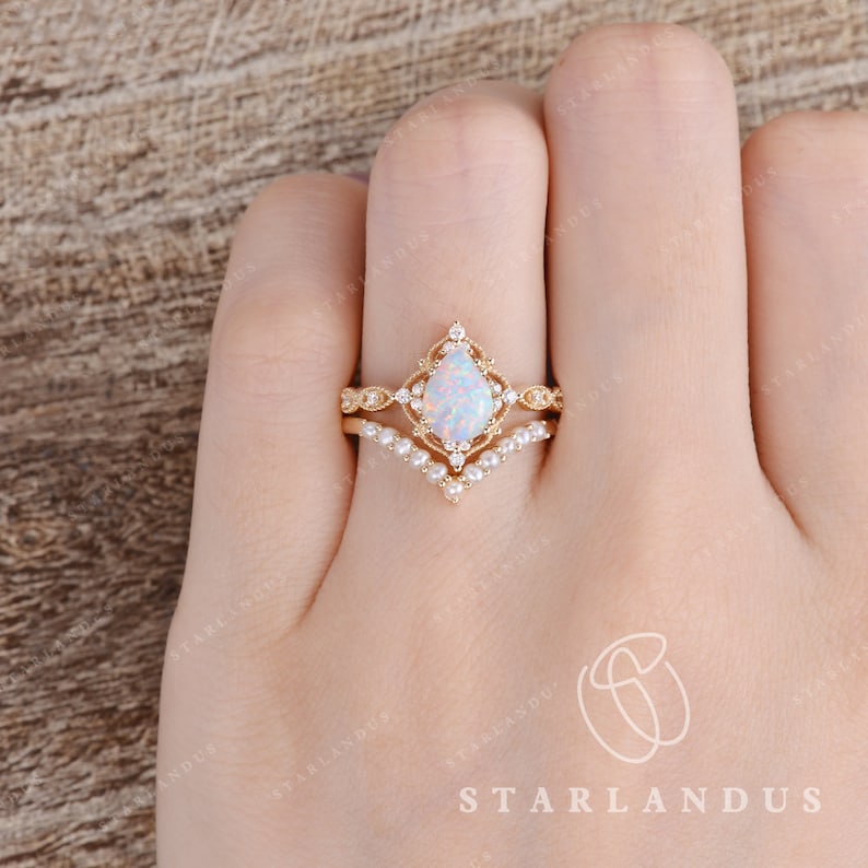 May include: A pear-shaped opal engagement ring with a delicate diamond halo setting. The ring is set on a gold band and is paired with a matching wedding band featuring a row of pearls.