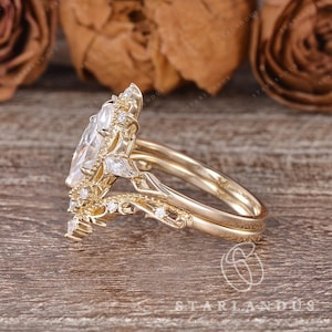 Puede incluir: Un anillo de oro con una gran piedra central de corte marquesa rodeada por un halo de piedras redondas y en forma de pera. La banda presenta un intrincado trabajo de metal en filigrana y piedras peque&ntilde;as a lo largo del aro.