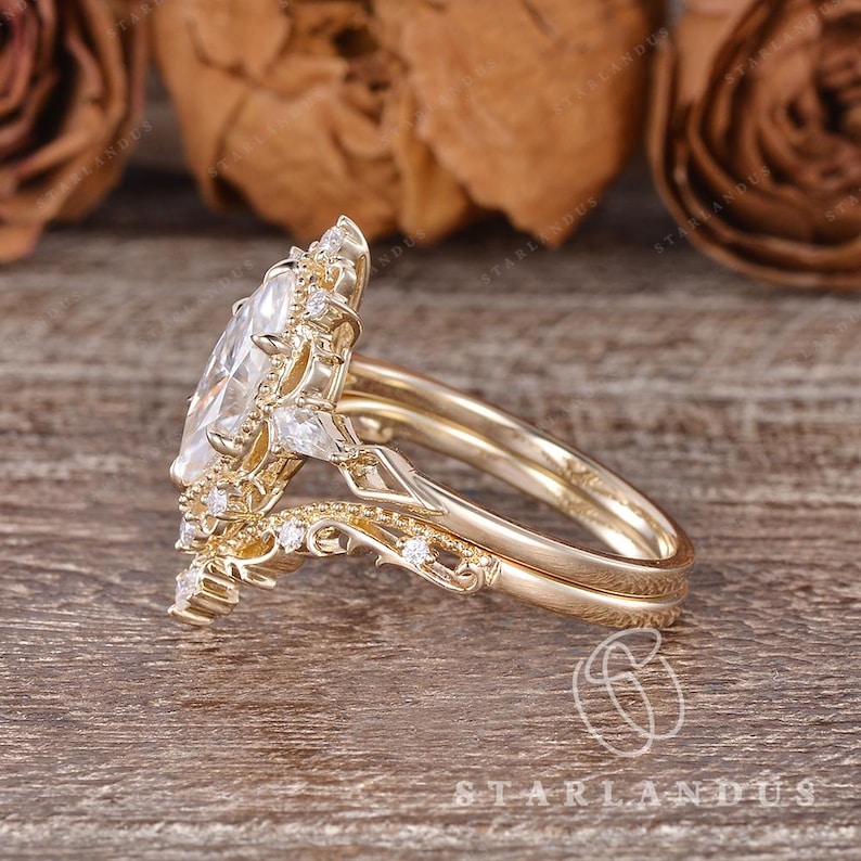 Puede incluir: Un anillo de oro con una gran piedra central de corte marquesa rodeada por un halo de piedras redondas y en forma de pera. La banda presenta un intrincado trabajo de metal en filigrana y piedras peque&ntilde;as a lo largo del aro.