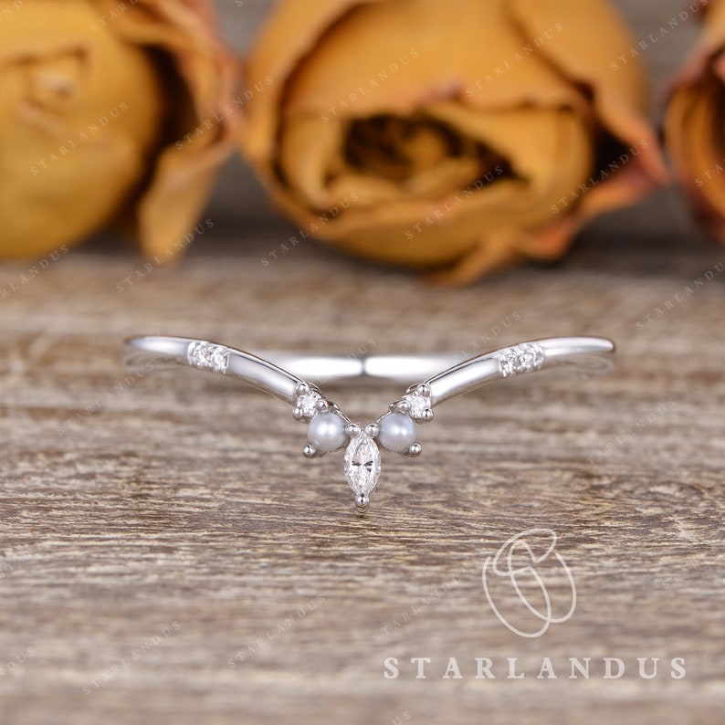 May include: A delicate silver ring with a V-shaped design. The ring is adorned with small diamonds and two pearl accents. The ring is set against a backdrop of brown wood.