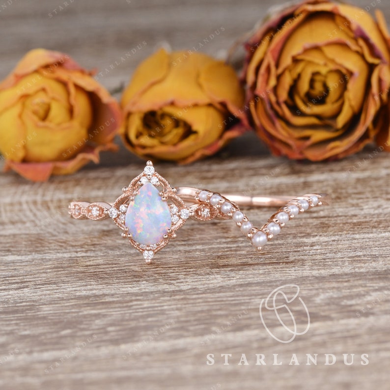 May include: A pear-shaped opal engagement ring set in rose gold with a delicate diamond halo and a matching pearled wedding band. The ring is set against a wooden background with dried roses.