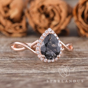 May include: A rose gold engagement ring with a pear-shaped black diamond center stone surrounded by a halo of smaller diamonds. The band is a twisted design.
