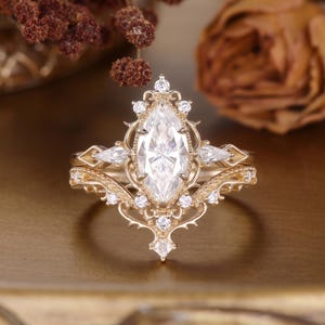 Puede incluir: Un conjunto de anillos de oro con una piedra central de corte marquesa rodeada de piedras redondas y en forma de pera. La banda de metal muestra detalles de filigrana intrincados y bordes puntiagudos, creando un aspecto ornamentado.