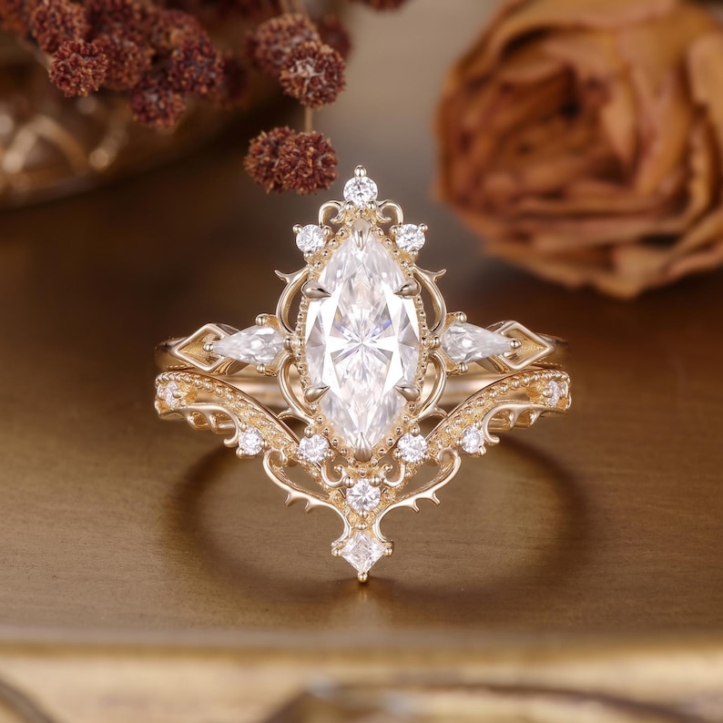 Puede incluir: Un conjunto de anillos de oro con una piedra central de corte marquesa rodeada de piedras redondas y en forma de pera. La banda de metal muestra detalles de filigrana intrincados y bordes puntiagudos, creando un aspecto ornamentado.