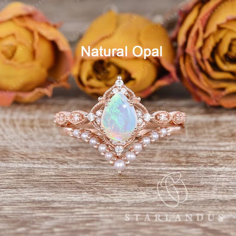 May include: A pear-shaped opal engagement ring set in rose gold with a delicate diamond halo and a pearl band. The ring is set on a wooden surface with dried flowers in the background. The text "Natural Opal" is displayed above the ring.