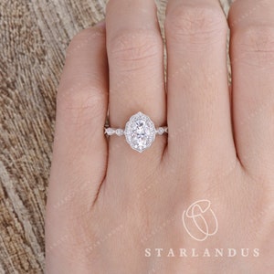 May include: A silver engagement ring with an oval-shaped center stone surrounded by smaller diamonds. The ring has a delicate, floral design and is set on a thin band.