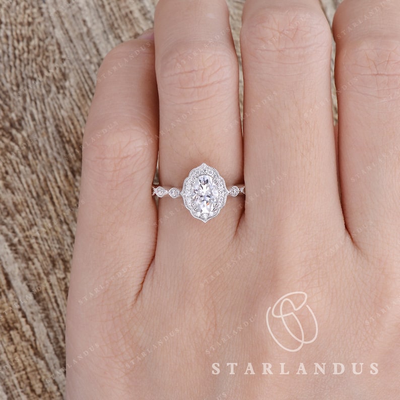 May include: A silver engagement ring with an oval-shaped center stone surrounded by smaller diamonds. The ring has a delicate, floral design and is set on a thin band.