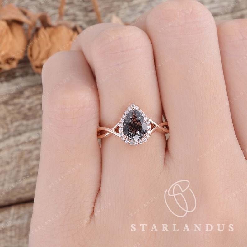 May include: A rose gold engagement ring with a pear-shaped black diamond center stone and a halo of smaller diamonds. The band is twisted and features a delicate design.