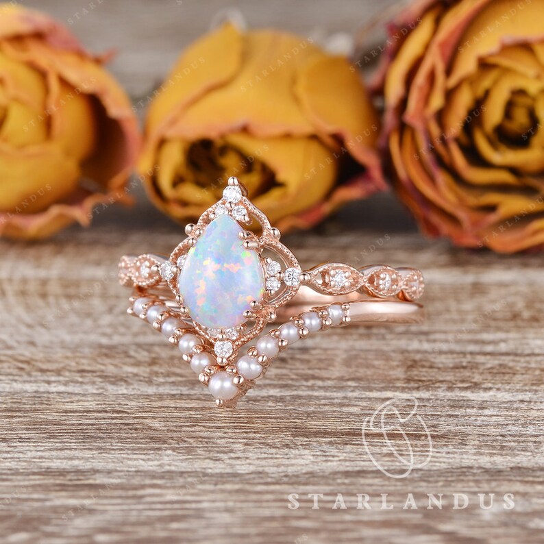May include: A pear-shaped opal engagement ring set in rose gold with a delicate diamond and pearl band. The ring features a vintage-inspired design with intricate details.