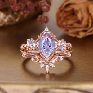 May include: A rose gold ring set featuring a pear-shaped, faceted purple-blue center stone surrounded by small clear white accent stones and two small light blue stones. The metal band has an intricate, ornate design with leaf-like details.