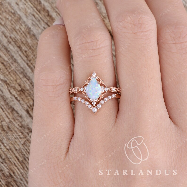 May include: A pear-shaped opal engagement ring set in rose gold with a delicate floral design and a matching band with small pearls. The ring is on a finger.